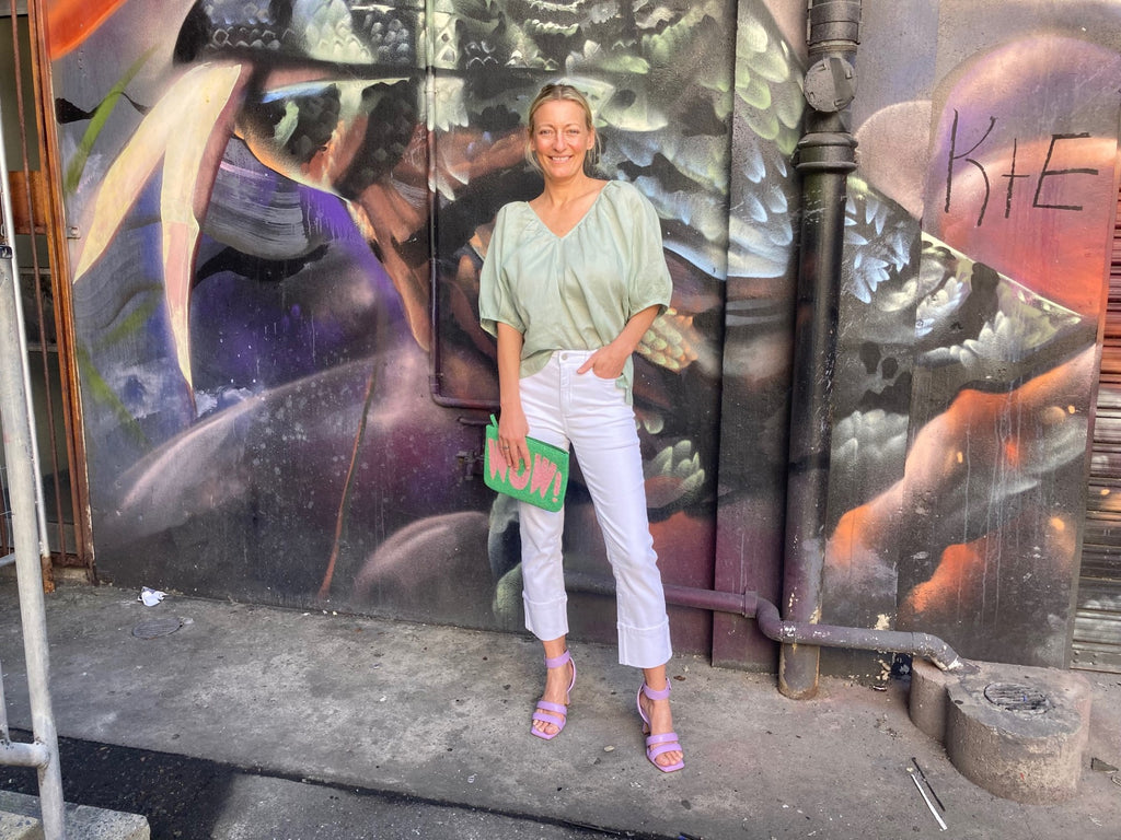 Model stands in front of graffiti wall wearing white Zoe Kratzmann jeans and green linen shirt, purple Cerrado heels and a green and purple beaded clutch purse